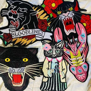Könnte beinhalten: Gestickte Aufnäher mit verschiedenen Designs. Enthält einen schwarzen Panther mit dem Wort "BLOODLINES", einen brüllenden Panther mit roten Rosen, einen schwarzen Pantherkopf, eine Katze in einem Mantel und einen rosa Hummer.