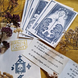 Op de afbeelding: Een set orakelkaarten met blauwe en witte illustraties en tekst. De kaarten bevatten afbeeldingen van heiligen en de woorden "STELLA MARIS" en "ORACLE-DES-SAINTS". De kaarten zijn gerangschikt op een gele ondergrond met gedroogde bloemen.