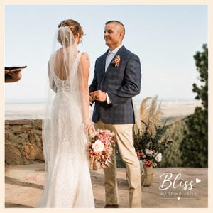 May include: A floor-length white lace wedding dress with a low back and a long sheer veil, paired with a navy blue plaid blazer, a white button-down shirt, and tan trousers. The wedding attire is worn by two individuals standing outdoors.