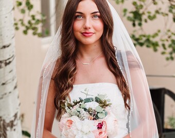 May include: A white bridal veil with delicate beaded trim worn over a strapless white lace dress, paired with a bouquet of pink and white peonies and eucalyptus. The text Bliss Veils AND ACCESSORIES is printed in green and grey on the image.