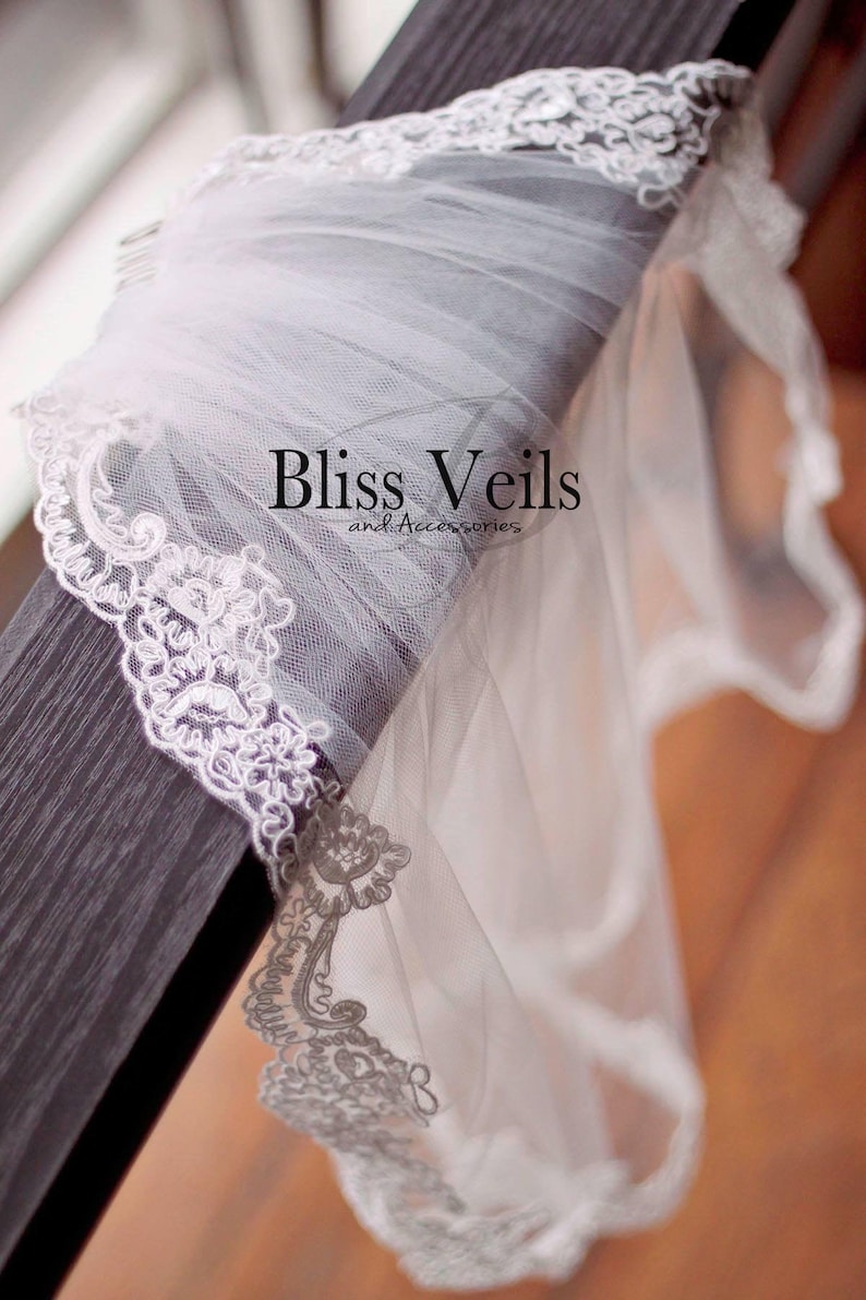 May include: A white bridal veil with a delicate lace trim. The veil is draped over a dark wooden surface. The text "Bliss Veils and Accessories" is visible in the center of the image.
