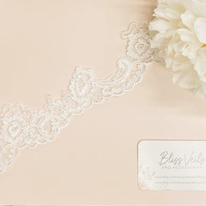 May include: White lace trim with a delicate floral pattern. The lace is shown on a light pink background with a business card that reads "Bliss Veils and Accessories".