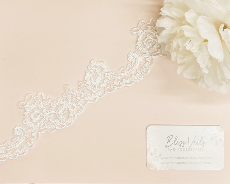 May include: White lace trim with a delicate floral pattern. The lace is shown on a light pink background with a business card that reads "Bliss Veils and Accessories".