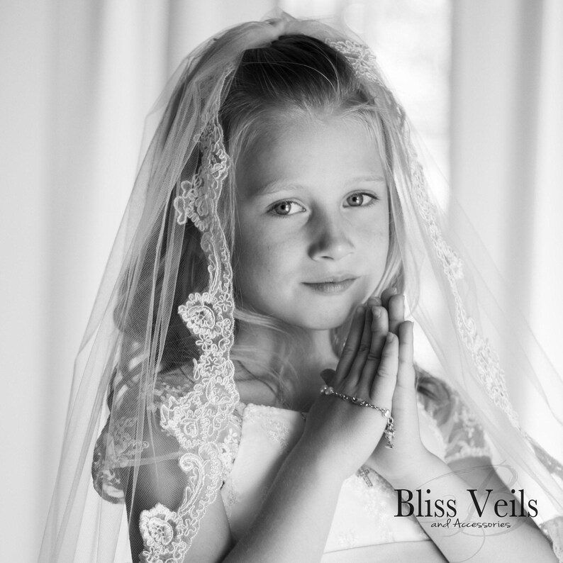 May include: A young girl wearing a white veil with lace trim, holding her hands together in prayer. The veil is made of soft, sheer fabric and has a delicate floral pattern. The girl is looking down at her hands, and her expression is serene.