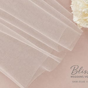 May include: A sheer, light blush-colored tulle veil with a raw edge. The fabric is folded in soft layers, creating a delicate texture. In the corner, the text reads: Bliss WEDDING VEILS &copy; RAW EDGE VEIL, accompanied by a small heart icon.