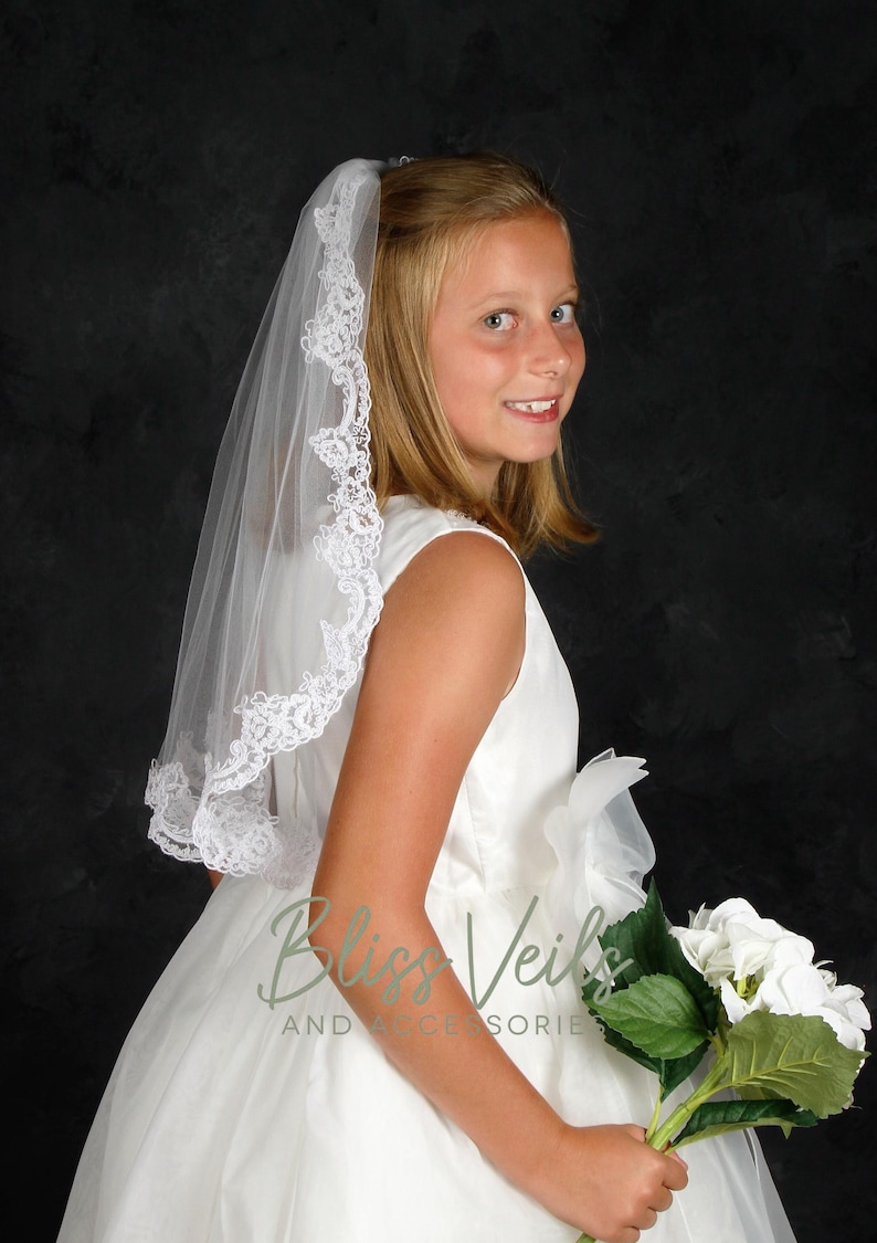 May include: A young person wearing a white dress and a white lace veil. They are holding a bouquet of white flowers.