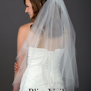 May include: A white, sheer, multi-layered tulle wedding veil attached to a hair comb. The veil features a soft, flowing texture and drapes over the back. The text Bliss Veils and Accessories is printed in black at the bottom of the image.