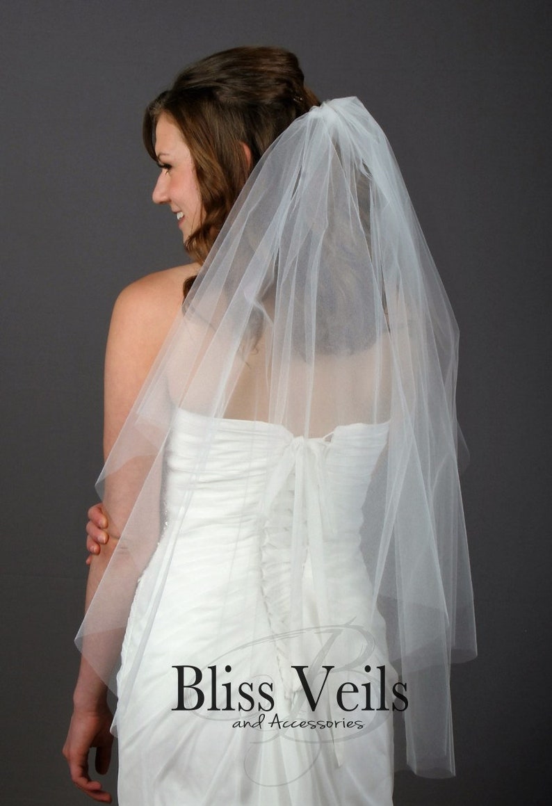 May include: A white, sheer, multi-layered tulle wedding veil attached to a hair comb. The veil features a soft, flowing texture and drapes over the back. The text Bliss Veils and Accessories is printed in black at the bottom of the image.
