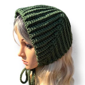 May include: A green knitted bonnet with a ribbed texture. The bonnet has a rounded shape and ties under the chin. The item is displayed on a mannequin head with blonde hair, against a white background.