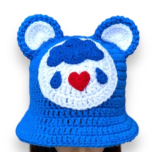 Puede incluir: Un sombrero de cubo de ganchillo azul hecho a mano con detalles en blanco y orejas de oso. El sombrero presenta un círculo blanco con un corazón rojo y gotas azules. El sombrero lo lleva una persona.