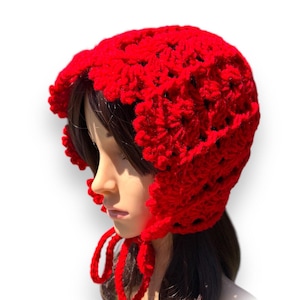 May include: A bright red, hand-crocheted bonnet featuring a scalloped trim and ear flaps. The bonnet is secured with long, red ties. The hat is displayed on a mannequin head against a white backdrop.