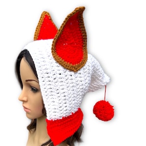 May include: A white crocheted hat with large red and brown-trimmed ears, a red neck piece, and a red pom-pom. The hat is designed to resemble a fox or other animal, and is likely a costume or novelty item.
