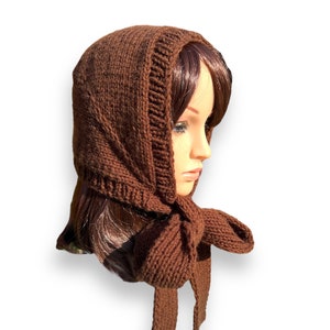 May include: A brown knitted hat with a long scarf attached. The hat is pulled down over the head and the scarf is tied in a bow at the neck.