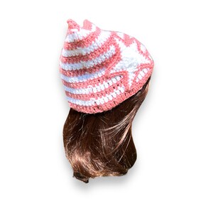 May include: A handmade crochet hat in a cat ear design. The hat features alternating stripes of white and a soft coral pink. The hat is shown on a person with brown hair. The hat is a unique and stylish accessory.