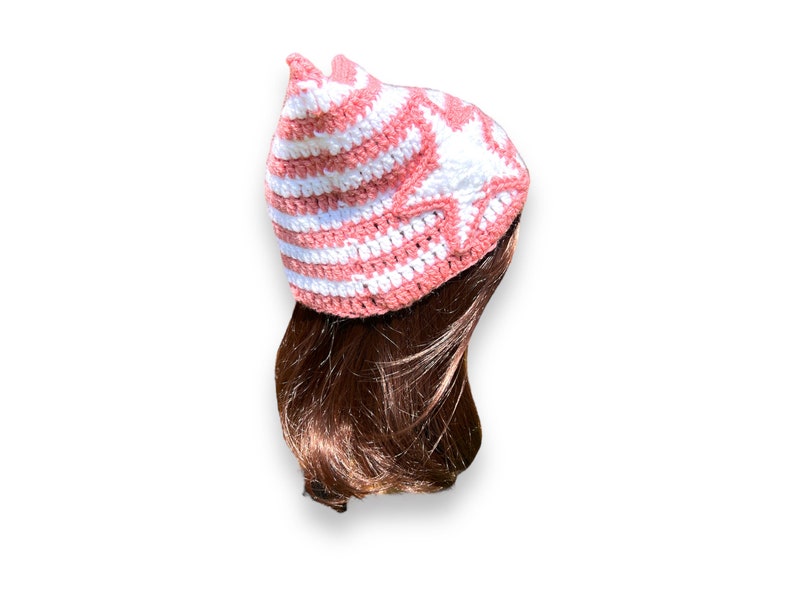 May include: A handmade crochet hat in a cat ear design. The hat features alternating stripes of white and a soft coral pink. The hat is shown on a person with brown hair. The hat is a unique and stylish accessory.