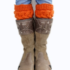 May include: A pair of brown leather boots with a decorative flower pattern on the side. The boots are worn with orange knitted leg warmers and blue jeans.