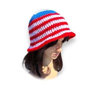 May include: A red, white, and blue crochet bucket hat. The hat features alternating red and white stripes with a blue crown and a red brim. The hat is handmade and has a classic, casual style.