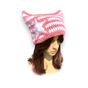 May include: A pink and white crocheted hat with pointed ears and a white star design on the front. The hat is made of yarn and is designed to be worn on the head. The hat is a unique and stylish accessory.