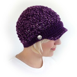 May include: Purple crocheted hat with a brim and a silver button. The hat is made with a loose stitch pattern.