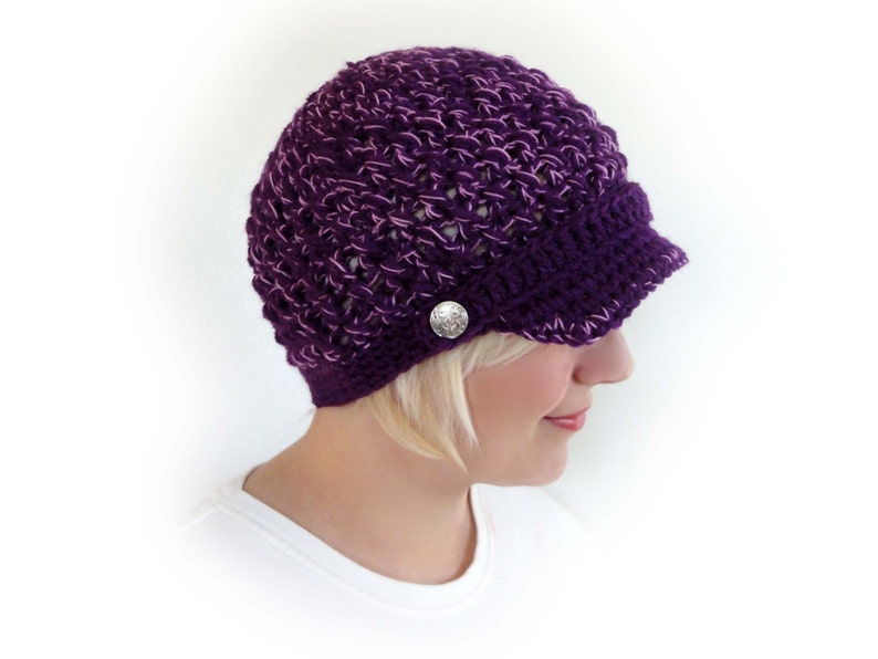 May include: Purple crocheted hat with a brim and a silver button. The hat is made with a loose stitch pattern.