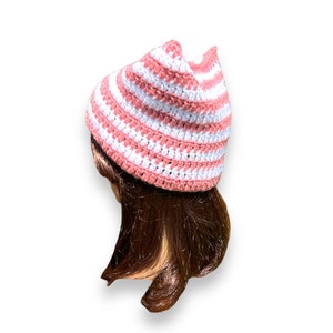 May include: A handmade crochet hat in a striped pattern of white and light pink. The hat has a pointed crown and is shown on a mannequin head with brown hair. The hat is a stylish accessory.