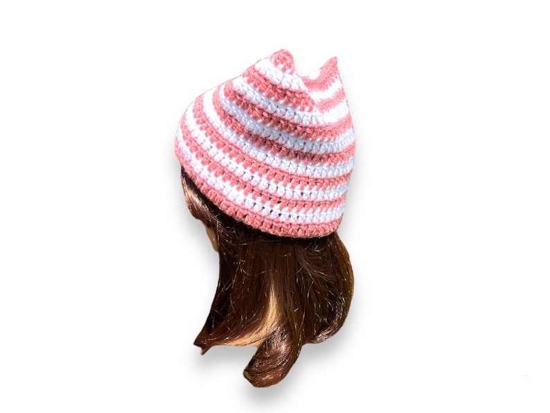 May include: A handmade crochet hat in a striped pattern of white and light pink. The hat has a pointed crown and is shown on a mannequin head with brown hair. The hat is a stylish accessory.