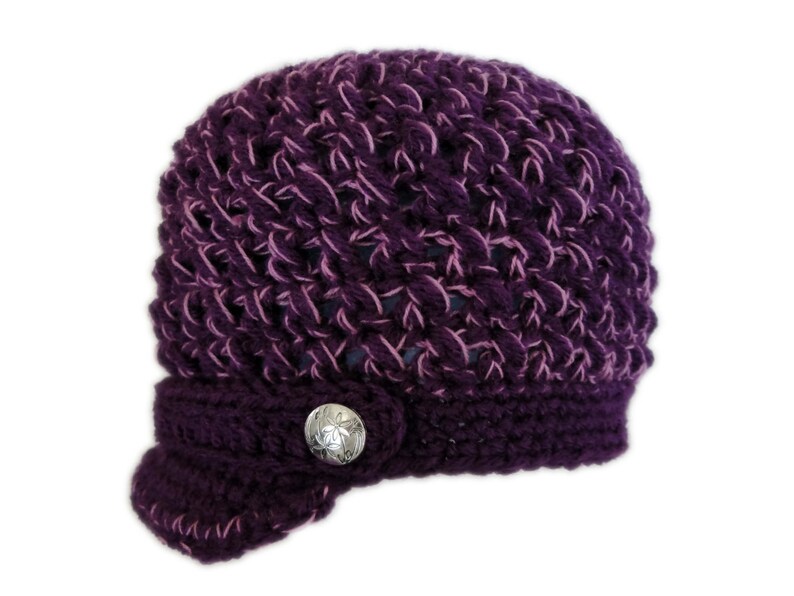 May include: A purple crocheted hat with a pink yarn detail and a silver button. The hat has a brim that is folded down on one side.