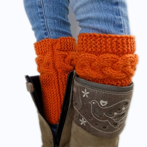 May include: A pair of orange knitted leg warmers with a cable knit pattern. The leg warmers are worn over a pair of blue jeans and brown boots.