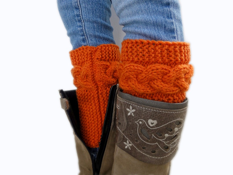 May include: A pair of orange knitted leg warmers with a cable knit pattern. The leg warmers are worn over a pair of blue jeans and brown boots.