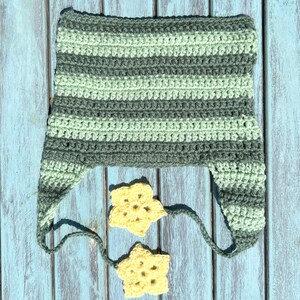 May include: A striped crocheted hat in shades of green with two yellow crocheted stars attached to the hat's ties. The hat and stars are lying on a blue wooden surface.