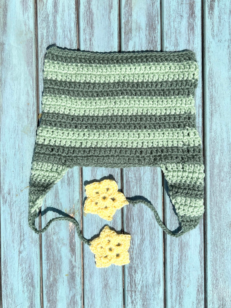 May include: A striped crocheted hat in shades of green with two yellow crocheted stars attached to the hat's ties. The hat and stars are lying on a blue wooden surface.