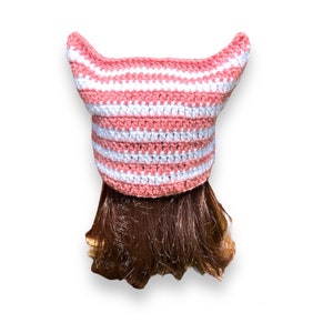 May include: A handmade crochet hat in a cat ear design. The hat features alternating horizontal stripes of white and light pink. The hat is shown on a person with brown hair, against a white background.