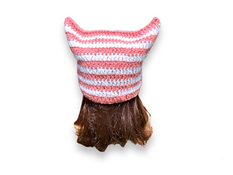 May include: A handmade crochet hat in a cat ear design. The hat features alternating horizontal stripes of white and light pink. The hat is shown on a person with brown hair, against a white background.