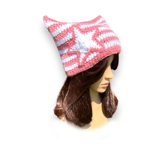 May include: A pink and white crocheted hat with a star design. The hat is made of yarn and has a pointed top. The hat is worn by a mannequin with brown hair.