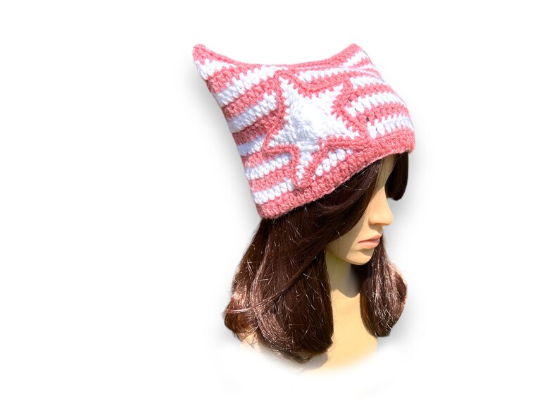 May include: A pink and white crocheted hat with a star design. The hat is made of yarn and has a pointed top. The hat is worn by a mannequin with brown hair.