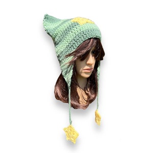 May include: A green and yellow crocheted hat with ear flaps and star-shaped tassels. The hat is made with a textured stitch and has a star design on the top.