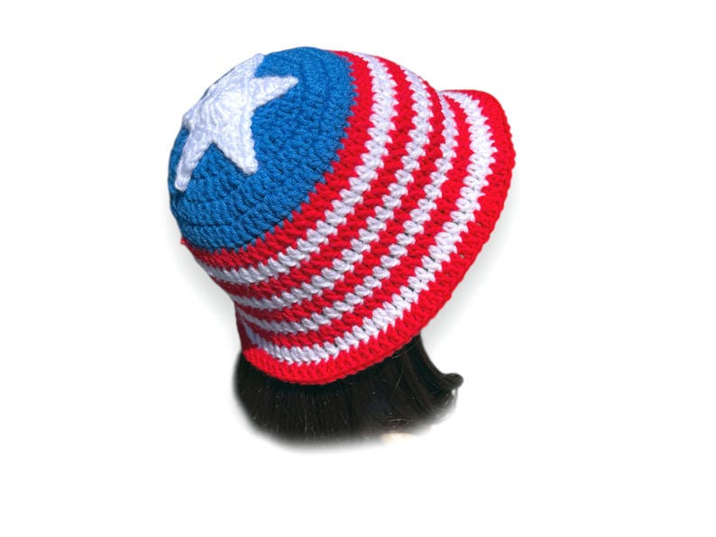 May include: A crocheted hat in red, white, and blue, featuring a white star on the blue crown. The hat has a brim and is designed with alternating red and white stripes, evoking a patriotic theme.