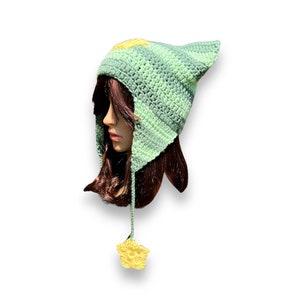 May include: A green and yellow crocheted hat with a pointed ear and a star-shaped tassel. The hat is made with a striped pattern and has a soft, cozy texture.