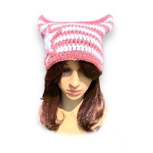 May include: A handmade crochet hat in pink and white stripes, featuring pointed cat ears. The hat is designed with a striped pattern and is likely made of soft yarn, perfect for a whimsical accessory.