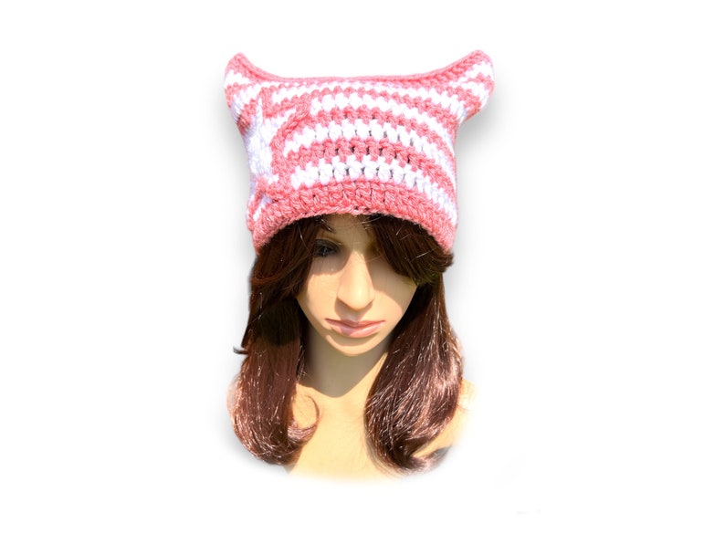 May include: A handmade crochet hat in pink and white stripes, featuring pointed cat ears. The hat is designed with a striped pattern and is likely made of soft yarn, perfect for a whimsical accessory.