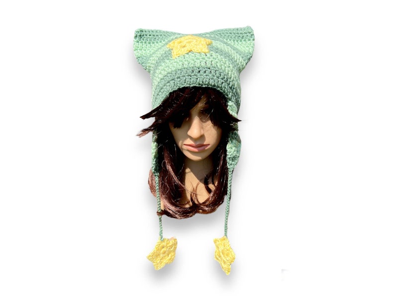 May include: A green and yellow crocheted hat with cat ears and star details. The hat has long ties with star ends.
