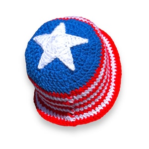 May include: A handmade crochet bucket hat in red, white, and blue, featuring a white star on a blue crown. The hat's brim has alternating red and white stripes, evoking a patriotic theme.
