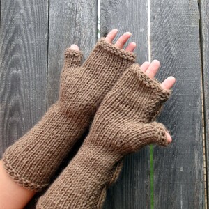 Puede incluir: Un par de guantes sin dedos de punto marr&oacute;n con textura acanalada. Los guantes se muestran sobre un fondo de madera.