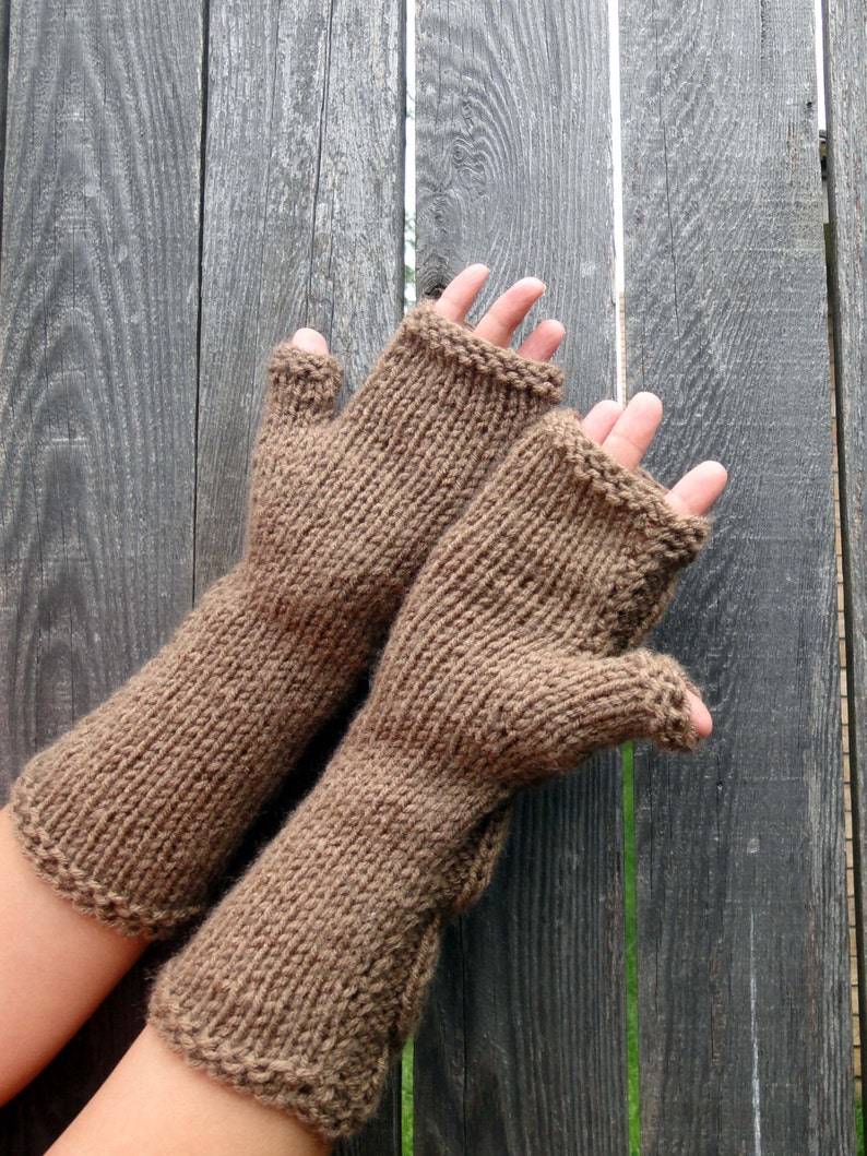 Puede incluir: Un par de guantes sin dedos de punto marr&oacute;n con textura acanalada. Los guantes se muestran sobre un fondo de madera.