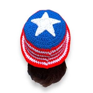 May include: A handmade crochet hat in red, white, and blue, featuring a white star on top. The hat has a brim with alternating red and white stripes. The hat is shown from a top-down perspective.