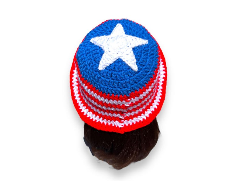 May include: A handmade crochet hat in red, white, and blue, featuring a white star on top. The hat has a brim with alternating red and white stripes. The hat is shown from a top-down perspective.