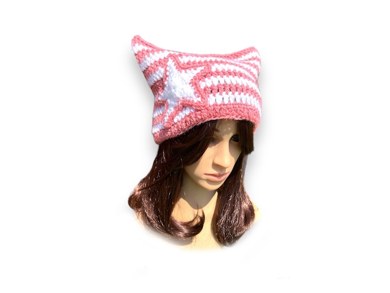 May include: A pink and white striped crochet hat with pointed cat ears. The hat features a star design on the front and is made of a soft, textured yarn. The hat is a unique and stylish accessory.