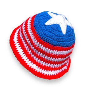 May include: A handmade crochet bucket hat in red, white, and blue. The hat features alternating red and white stripes on the brim and a blue crown with a white star. This patriotic hat is perfect for summer.