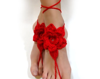 May include: Red crochet barefoot sandals with a floral design. The sandals feature a large flower on the top of the foot and straps that wrap around the ankle. The sandals are made of red yarn.