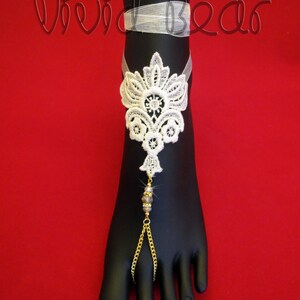 May include: White lace foot jewelry with a gold chain and crystal beads. The jewelry is on a black foot model against a red background.